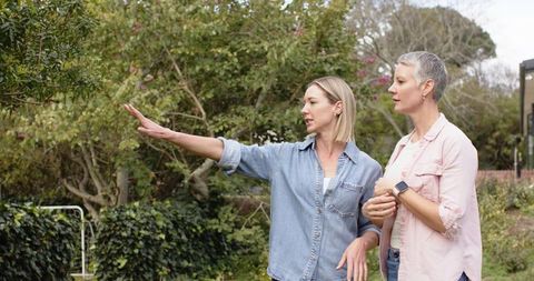Two women discussing garden plans pointing at plants in backyard casual conversation