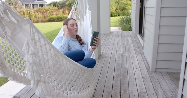 Young woman relaxing on a hammock while texting on her smartphone. The setting is a wooden porch with greenery in the background. Useful for concepts related to leisure, casual moments, remote communication, outdoor lifestyle, and relaxation.