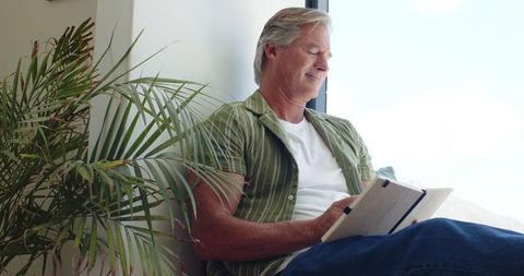 Senior man journaling on sunlit window seat with indoor palm and cozy minimalist nook