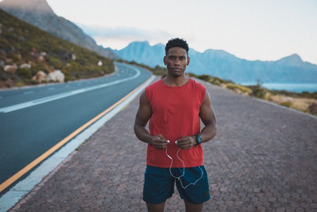 African American man in athletic wear standing on a coastal road holding headphones. Ideal for promoting healthy lifestyles, fitness training programs, outdoor activities, and athletic apparel. Suitable for use in advertisements, fitness blogs, and health-related articles.