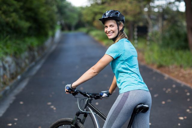 Woman wearing helmet and athletic wear, standing with mountain bike on a paved road surrounded by nature. Ideal for promoting outdoor activities, fitness, healthy lifestyle, and cycling adventures.
