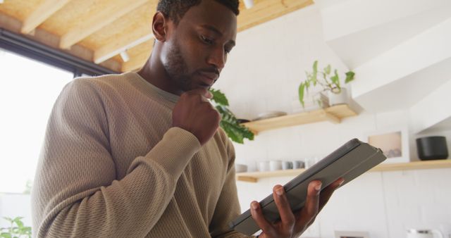 A man is reading on a tablet while standing in a modern kitchen. This image depicts a casual, relaxed atmosphere and represents the integration of technology into everyday home life. It can be used for topics related to digital lifestyles, home technology, or modern living.