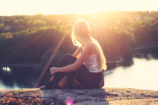 Girl sits on cliff edge at sunset, with sun rays creating a serene atmosphere. Perfect for travel, adventure, and relaxation themes, emphasizing connection with nature and peaceful contemplation. Ideal for blogs, advertisements, and social media focusing on travel, self-reflection, or natural beauty.