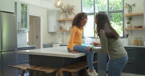 Asian mother handing green cup to african american child on kitchen island, caregiving moment