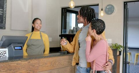 Smiling barista greeting diverse couple making contactless payment at modern coffee counter