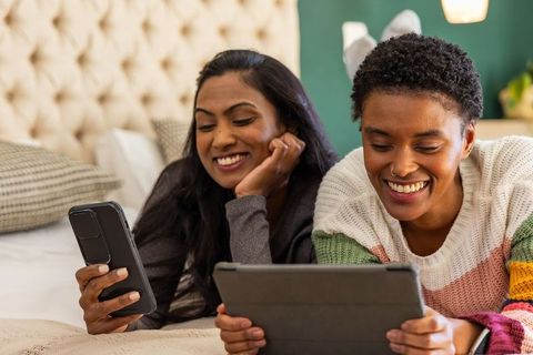 Smiling indian and african american couple relaxing on bed using tablet and smartphone