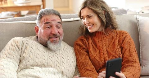 Couple relaxing on sofa wearing cozy cable-knit sweaters sharing smartphone