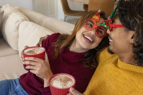 Festive couple sharing cozy cocoa on sofa wearing holiday glasses and showing ring