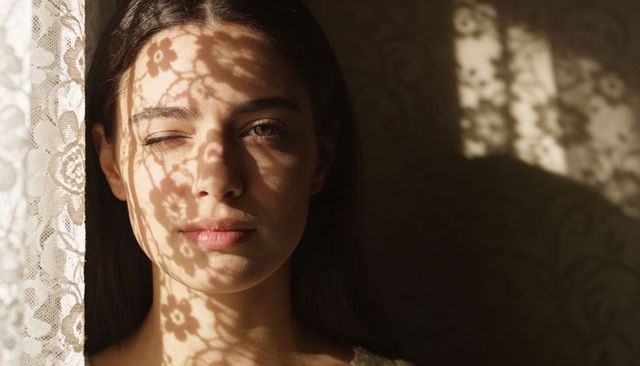 Young woman standing by window wearing light top casting lace floral shadows on face