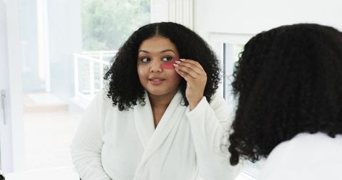 African american woman applying under-eye gel patch at vanity mirror for skincare routine