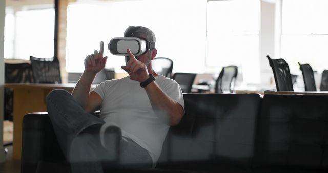 Man wearing VR headset sitting on couch in modern office environment, interacting with invisible interface. Can be used for themes related to cutting-edge technology, workplace innovation, virtual experiences, and futuristic corporate settings.