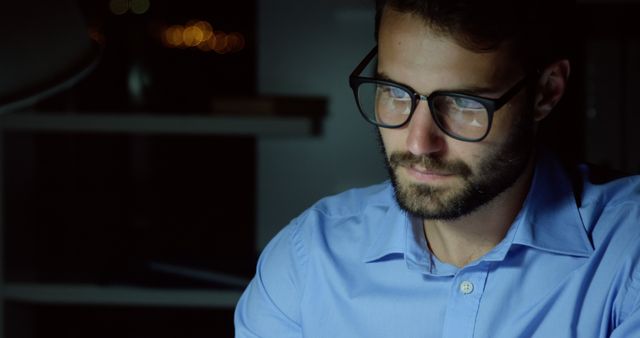 This image depicts a man with a beard and glasses, wearing a blue shirt, working late in an office. He appears thoughtful and focused, illuminated by soft lighting in a dark surrounding. Ideal for concepts like dedication, late hours, work ethic, productivity, or professional life.