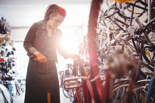 Female mechanic pumping air into a bicycle tire in a workshop filled with various bikes and tools. Ideal for use in articles about bike maintenance, cycling, women's roles in mechanical professions, or promoting bicycle repair services.