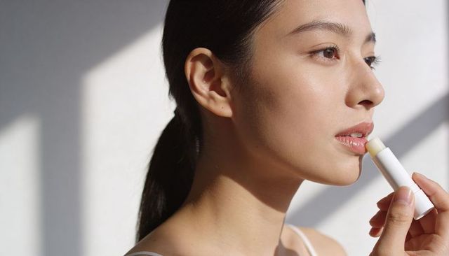 Asian woman applying lip balm in soft window light for natural dewy skincare beauty