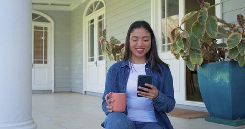 Young woman relaxing on veranda steps holding pink mug and checking smartphone