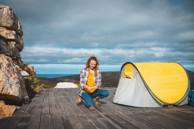 Caucasian woman setting up her tent on a wooden deck in a mountainous area. Ideal for use in travel blogs, adventure magazines, camping gear advertisements, and lifestyle articles focusing on outdoor activities and nature exploration.