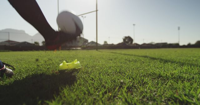 Image represents close-up view of rugby player's foot kicking a rugby ball on green grass field with goalpost in background. Ideal for use in sports-related content, advertising for sports footwear or equipment, and articles focused on rugby matches or athletic skills.