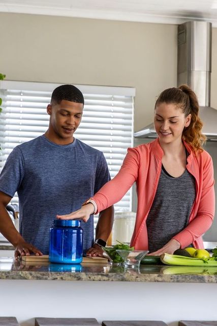 Couple preparing healthy smoothie mix at kitchen counter reaching for blue supplement jar