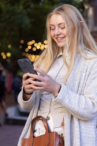 Smiling young woman texting on smartphone at dusk in cardigan, earbuds and tan handbag