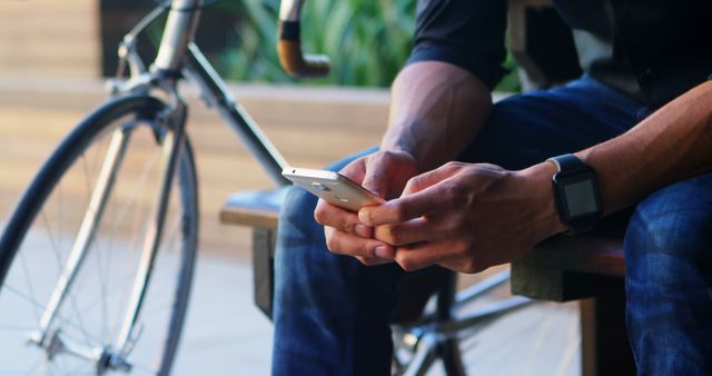 Man sits on bench texting on smartphone next to a bicycle outdoors, ideal for concepts of connectivity, leisure, and urban lifestyle. Suitable for use in technology promotions, lifestyle blogs, or advertisements highlighting casual use of mobile devices.