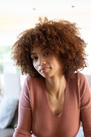African american woman sitting on sofa in sunlit living room wearing terracotta ribbed sweater