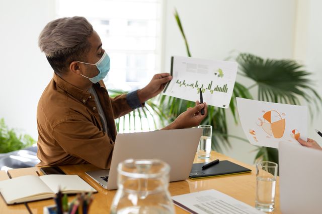 Businessman wearing a mask presenting graphs to colleagues during an office meeting. Ideal for use in articles about workplace safety, business presentations, teamwork, and professional environments during the pandemic.