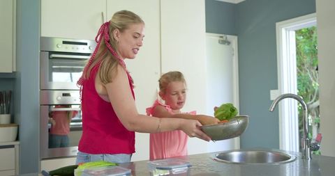 Mother and daughter washing lettuce at kitchen island, bright modern family cooking moment