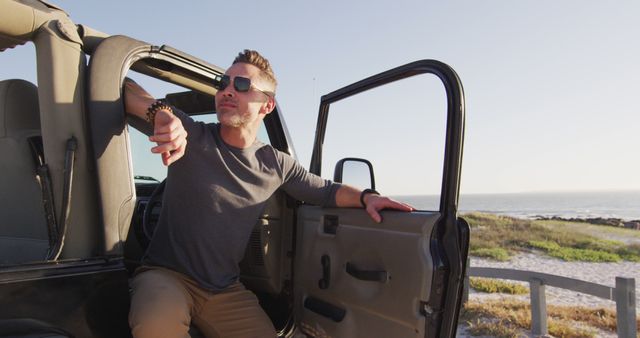 Man wearing sunglasses leaning on SUV door, enjoying serene coastal scenery. Ideal for travel, adventure, and lifestyle concepts promoting outdoors and freedom.
