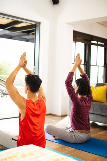 Rear view of gay multiracial couple with arms raised meditating while sitting on mats at home. Copy space, unaltered, love, together, homosexual, exercise, zen, yoga, fitness and active lifestyle.