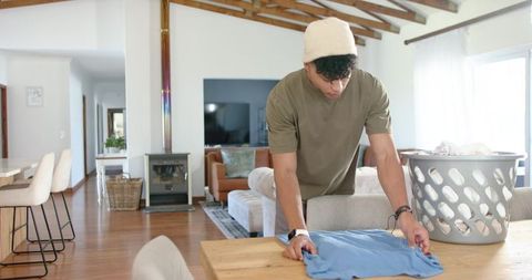 African american man folding laundry on wooden table in modern open-plan living room