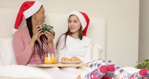 Women wearing santa hats sharing cozy breakfast in bed with gift and croissants