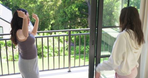 Two women stretching and talking on balcony as another opens sliding glass door to backyard
