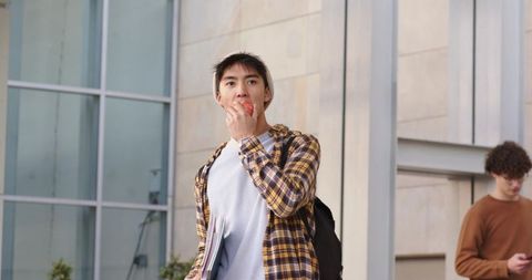 Asian student eating apple while walking on modern campus holding books and backpack