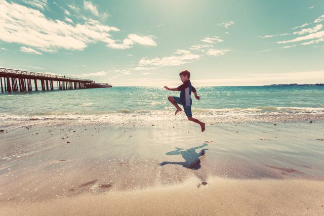 Child joyfully jumping on sandy beach near ocean waves under sunny sky. Ideal for ads related to travel, vacations, summer activities, family outings, and carefree childhood moments. Perfect for blog posts, social media content, or travel brochures highlighting beachside fun and summer enjoyment.