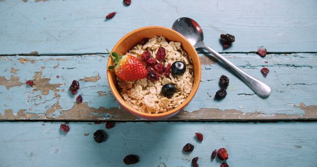 A bowl of cereal with fresh strawberries and blueberries sits on a rustic blue wooden table, with copy space. Scattered dried cranberries add a touch of color and texture to the wholesome breakfast scene.