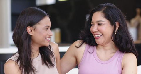 Two diverse women laughing together on couch in modern open-plan kitchen sharing warm moment