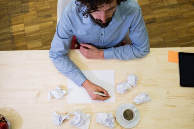Businessman sitting at desk writing on blank paper, surrounded by crumpled paper and a coffee cup. Ideal for illustrating concepts of brainstorming, creativity, office work, and professional tasks. Suitable for use in business presentations, articles on productivity, and creative process visuals.