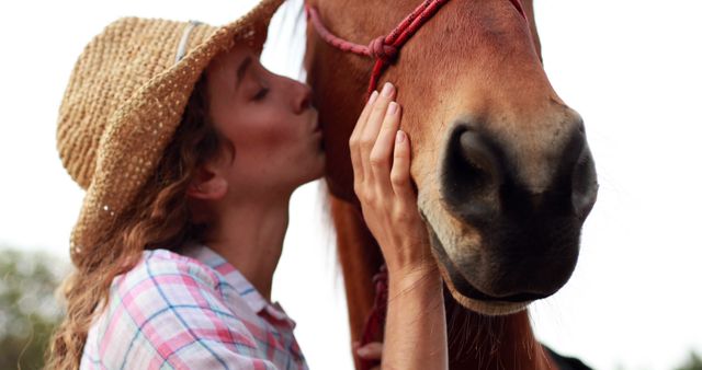 Side view of caucasian woman in straw hat kissing horse's head with copy space. Horse, horse riding, animal and love concept, unaltered.