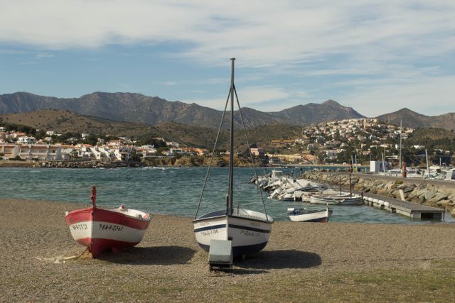 Small boats resting on a beach near a marina in a seaside village. Mountains are visible in the background under a cloudy sky. Perfect for travel articles, coastal living blogs, and nautical-themed designs.