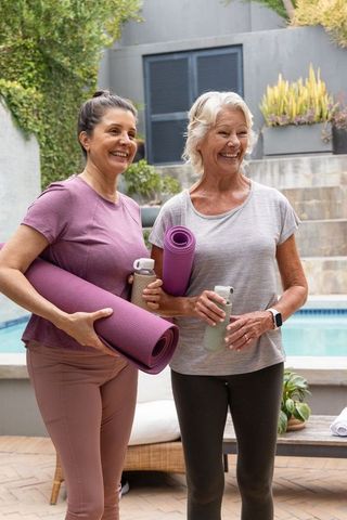 Mature women holding purple yoga mats and water bottles by poolside, smiling and chatting