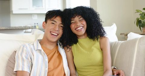 Joyful diverse couple laughing and leaning together on cozy modern bright living room sofa