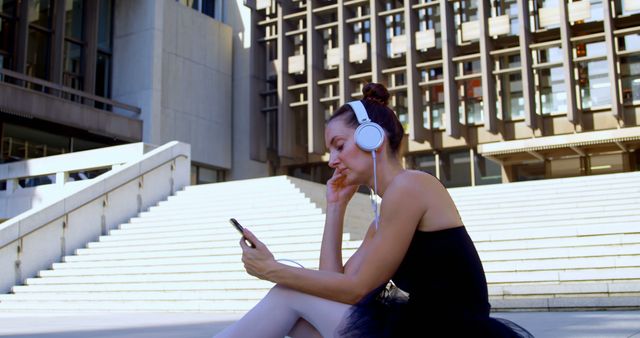 Ballerina in tutu relaxing on city stairs outside a modern building, listening to music on headphones while using a smartphone. Suitable for content related to dance, urban life, relaxation, and technology usage. Ideal for blogs, advertisements, and social media campaigns focusing on lifestyle and the arts.