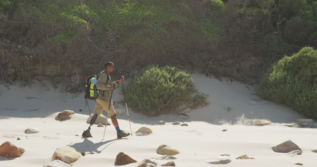 Man with prosthetic leg trekking through sandy and rocky beach landscape with backpack. Demonstrates resilience, adventure spirit, and love for nature. Ideal for use in outdoor activity promotions, motivational and inspirational stories, advertisements for hiking and camping gear.