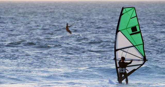 Windsurfer maneuvering across ocean waves with a green sail, while a kite surfer is seen in the distance. This dynamic and energetic activity is ideal for illustrating themes of adventure, outdoor sports, and water recreation. Perfect for travel brochures, outdoor sports blogs, and magazines featuring summer activities.