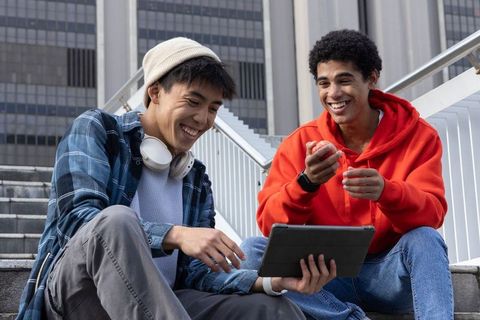 Teen friends laughing and sharing tablet on urban stairs wearing hoodie and beanie