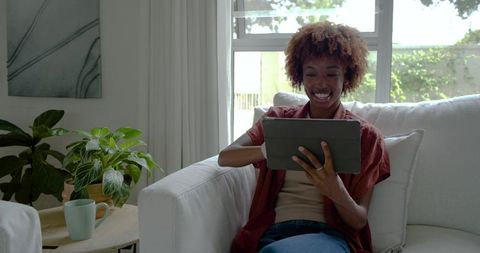 African american woman relaxing on sofa using tablet and smiling in sunlit living room