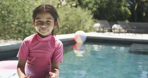 Smiling girl holding sunglasses on pink float by pool deck, summer backyard relaxation