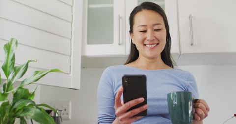 Smiling Woman Enjoys Coffee Break While Browsing Phone at Home