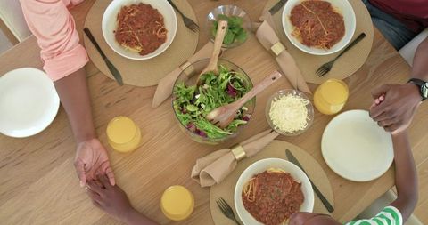 African american family holding hands around dinner table sharing spaghetti and salad