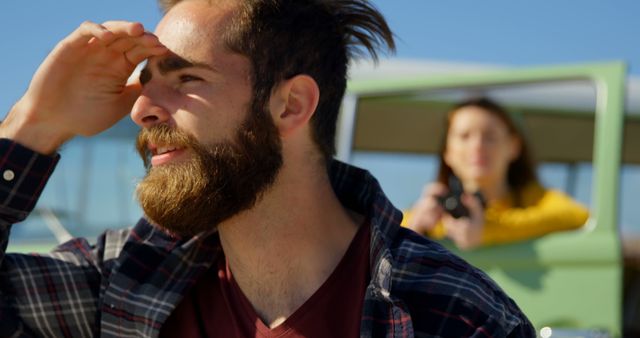 Young man with a beard and a plaid shirt is looking into the distance, enjoying a sunny day. Behind him, a woman in a yellow shirt is observing from a car, indicating a possible road trip or travel adventure. Ideal for themes related to outdoor activities, adventure, friendship, travel, and lifestyle.